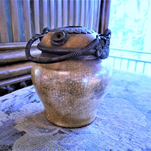May include: A small, cream-colored ceramic jar with a crackled glaze. The jar has a metal lid with a decorative handle and a dragon-shaped finial.