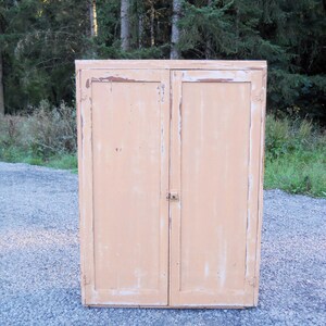 May include: A vintage, two-door cabinet with a light peach paint finish. The cabinet has a distressed look with some of the paint chipped away, revealing the wood underneath. The cabinet has two doors with simple, brass hinges and a small, brass latch.