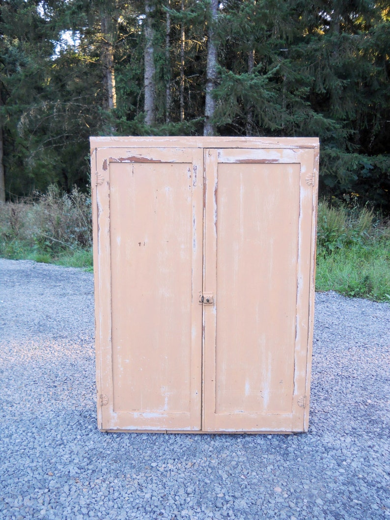 May include: A vintage, two-door cabinet with a light peach paint finish. The cabinet has a distressed look with some of the paint chipped away, revealing the wood underneath. The cabinet has two doors with simple, brass hinges and a small, brass latch.