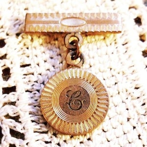 May include: Gold-toned brooch with a round pendant featuring a raised letter 'C' in a decorative circle. The pendant is attached to a bar with a textured surface.