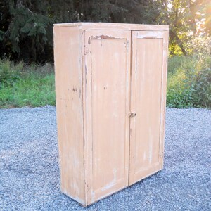 May include: A vintage, two-door cabinet with a light peach paint finish. The cabinet has a distressed look with chipped paint and visible wood grain. The cabinet is standing on a gravel surface.