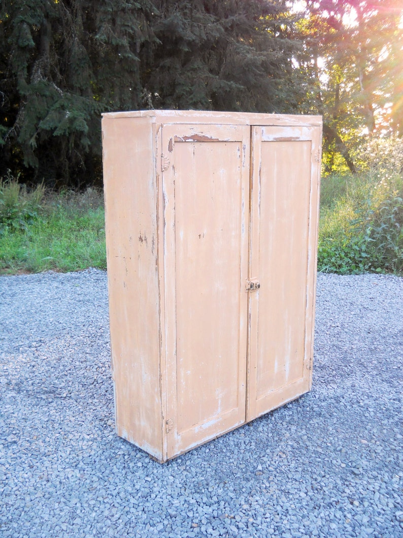 May include: A vintage, two-door cabinet with a light peach paint finish. The cabinet has a distressed look with chipped paint and visible wood grain. The cabinet is standing on a gravel surface.