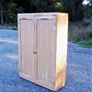 May include: A vintage wooden cabinet with two doors, painted in a light beige color with white paint chipping. The cabinet has a simple design with a flat top and a slightly rounded bottom.