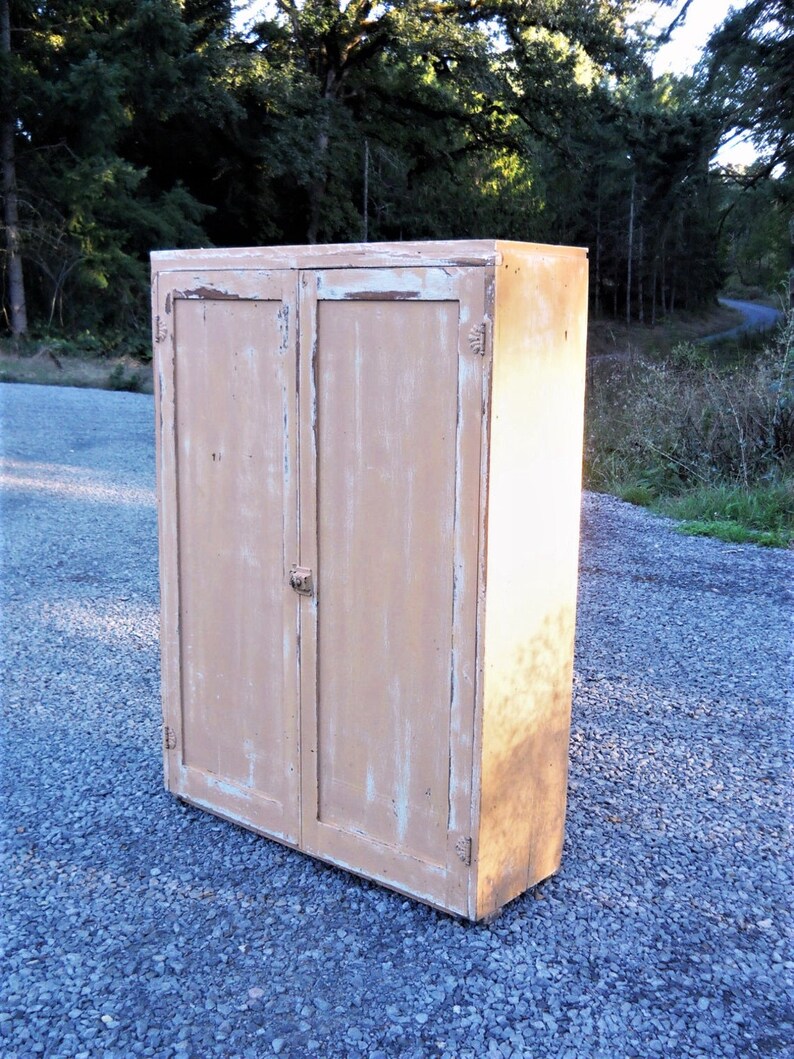 May include: A vintage wooden cabinet with two doors, painted in a light beige color with white paint chipping. The cabinet has a simple design with a flat top and a slightly rounded bottom.