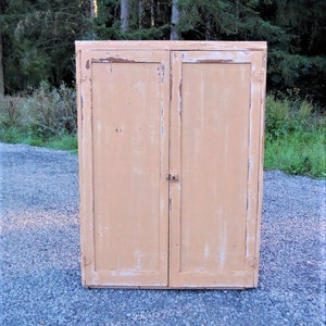 May include: A vintage, two-door cabinet with a distressed, light peach paint finish. The cabinet has a simple design with a single latch on the right door.
