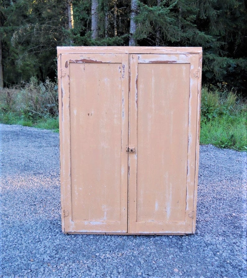 May include: A vintage, two-door cabinet with a distressed, light peach paint finish. The cabinet has a simple design with a single latch on the right door.