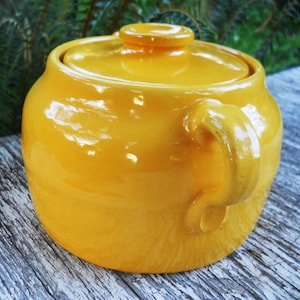 May include: A yellow ceramic crock pot with a lid and a handle. The crock pot is sitting on a wooden surface.