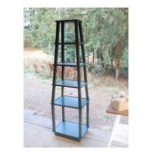 May include: A tall, dark green wooden shelf unit with five shelves. The shelf unit is standing on a concrete surface in front of a background of trees.