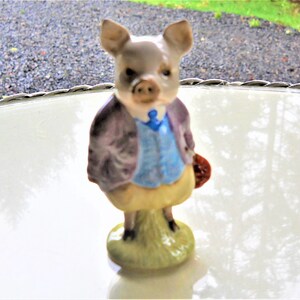 May include: A ceramic figurine of a pig wearing a purple jacket, blue vest, and yellow pants. The pig is holding a red book and standing on a green base.