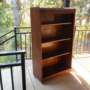 May include: A dark brown wooden bookcase with four shelves. The bookcase is standing on a concrete patio with a black metal railing in the background.