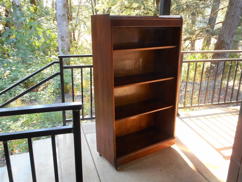 May include: A dark brown wooden bookcase with four shelves. The bookcase is standing on a concrete patio with a black metal railing in the background.