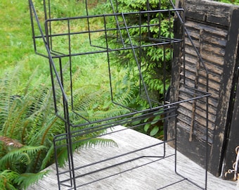 May include: Black metal magazine rack with three tiers. The rack is standing on a wooden surface.