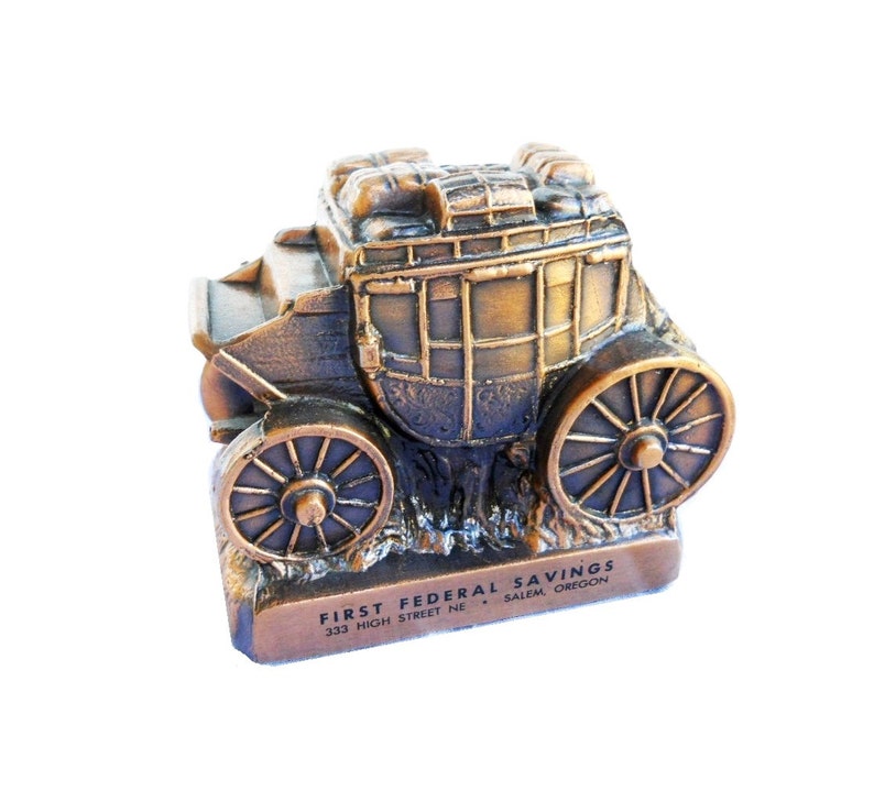May include: A bronze-coloured metal money box shaped like a stagecoach with wheels. The bank has the text "FIRST FEDERAL SAVINGS 333 HIGH STREET NE SALEM, OREGON" on the base.