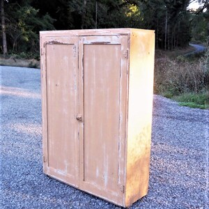 May include: A vintage, two-door cabinet with a light brown finish. The cabinet has a distressed look with chipped paint and visible wood grain. The cabinet is standing on a gravel surface.