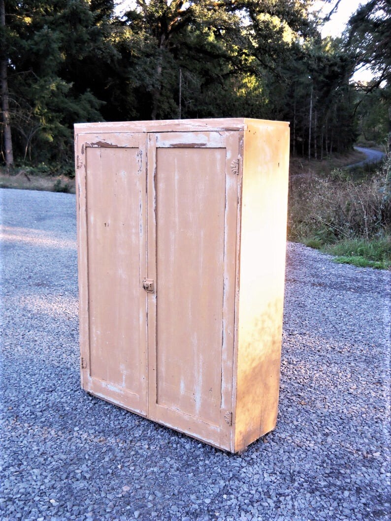 May include: A vintage, two-door cabinet with a light brown finish. The cabinet has a distressed look with chipped paint and visible wood grain. The cabinet is standing on a gravel surface.