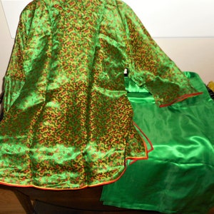 K&ouml;nnte beinhalten: Eine gr&uuml;ne Seidenrobe mit Blumenmuster und rotem und gelbem Besatz. Die Robe ist in der H&auml;lfte gefaltet und auf einem gr&uuml;nen Seidenstoff gelegt.