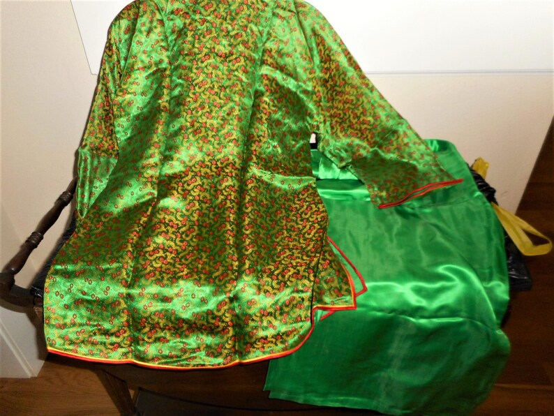 K&ouml;nnte beinhalten: Eine gr&uuml;ne Seidenrobe mit Blumenmuster und rotem und gelbem Besatz. Die Robe ist in der H&auml;lfte gefaltet und auf einem gr&uuml;nen Seidenstoff gelegt.