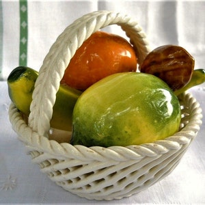 May include: A white ceramic basket with a woven design holds a collection of ceramic fruit. The fruit includes a yellow banana, a green lime, an orange, and a brown walnut.