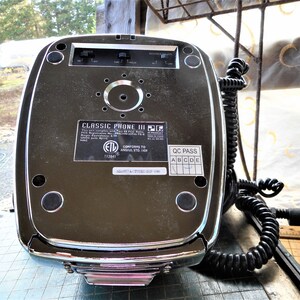 K&ouml;nnte beinhalten: Die R&uuml;ckseite eines silbernen Classic Phone III mit einem schwarzen Spiralkabel. Das Telefon hat ein schwarzes Etikett mit der Aufschrift "Classic Phone III" und enth&auml;lt die FCC-ID, das ETL-Zeichen und den Text "Conforms to ANSI Std. 101". Das Telefon hat auch ein schwarzes Etikett mit der Aufschrift "QC Pass" mit H&auml;kchen neben den Buchstaben A, B, C, D und E.