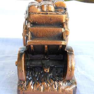 May include: A bronze-coloured metal piggy bank shaped like a covered wagon with wheels and a bed full of luggage. The wagon is on a base that looks like grass.