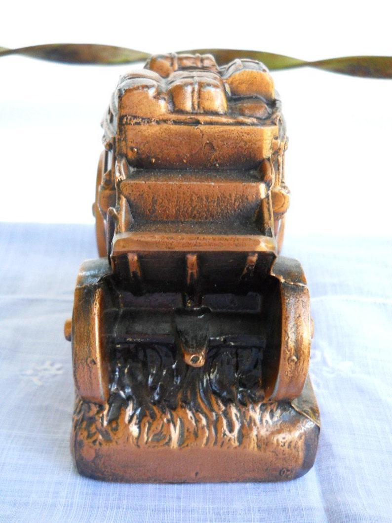 May include: A bronze-coloured metal piggy bank shaped like a covered wagon with wheels and a bed full of luggage. The wagon is on a base that looks like grass.