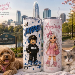 May include: Two cylindrical tumblers with clear straws. The left tumbler features a blue graffiti-style name JAYDEN, a cartoon character in a black Roblox hoodie, and blue stars. The right tumbler features the name Amina, a cartoon girl in a pink dress, and hearts.