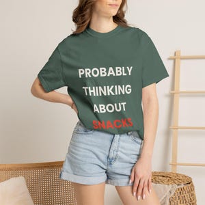 Pode incluir: Uma t-shirt verde escura com a frase "PROBABLY THINKING ABOUT SNACKS" em branco e vermelho. Combinada com calções de ganga. O conjunto tem um estilo casual.