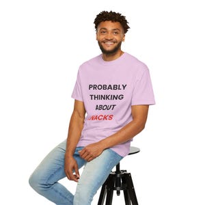 Pode incluir: Camiseta lilás claro com o texto "PROBABLY THINKING ABOUT SNACKS" em preto e vermelho. A pessoa está usando jeans azuis e sentada em um banquinho preto. A pessoa está sorrindo.