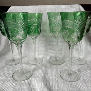 May include: A set of five green glass wine glasses featuring clear stems and bases. Each bowl displays a cut glass pattern with a cross-hatched design near the rim and etched grape clusters and leaves encircling the middle section of the glass.