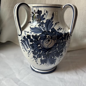 Peut inclure: Un vase en céramique blanche doté de deux anses, orné de motifs floraux peints à la main en bleu foncé. Le décor présente une rose centrale entourée de feuilles et de tiges, avec un liseré bleu sur le bord, les anses et la base.
