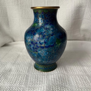 May include: A small cloisonné vase featuring a deep blue background with intricate floral patterns in shades of light blue, teal, and green. The design is outlined in thin gold metal wire, and the rim and base are finished with a smooth gold-toned metal edge.