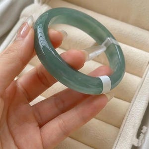 May include: A smooth, translucent, light green jade bangle bracelet with a rounded profile. The circular band features a polished, glossy surface and includes a small, white rectangular security tag attached to the side of the jade material.