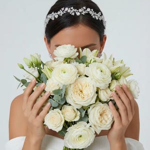 May include: A silver-toned headband featuring a delicate floral design with clear crystals and rhinestones, worn with a large bouquet of white roses, ranunculus, and eucalyptus greenery, paired with small silver crystal stud earrings.