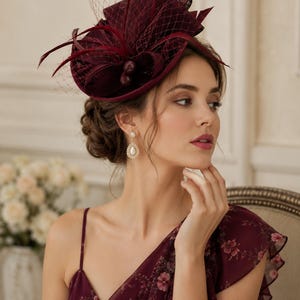 May include: A deep burgundy fascinator hat featuring a structured base, delicate netting, and feather accents. The headpiece is paired with gold-toned drop earrings featuring a central pearl surrounded by small clear stones and a floral dress.