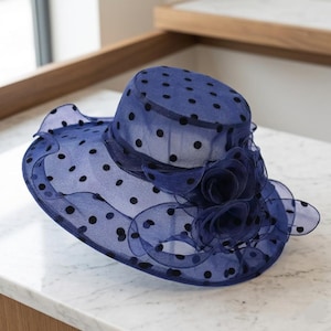 May include: A navy blue organza hat featuring a black polka dot pattern throughout the sheer fabric. The hat has a wide, wavy brim and is decorated with a cluster of layered, ruffled fabric flowers on the side of the crown and brim base.