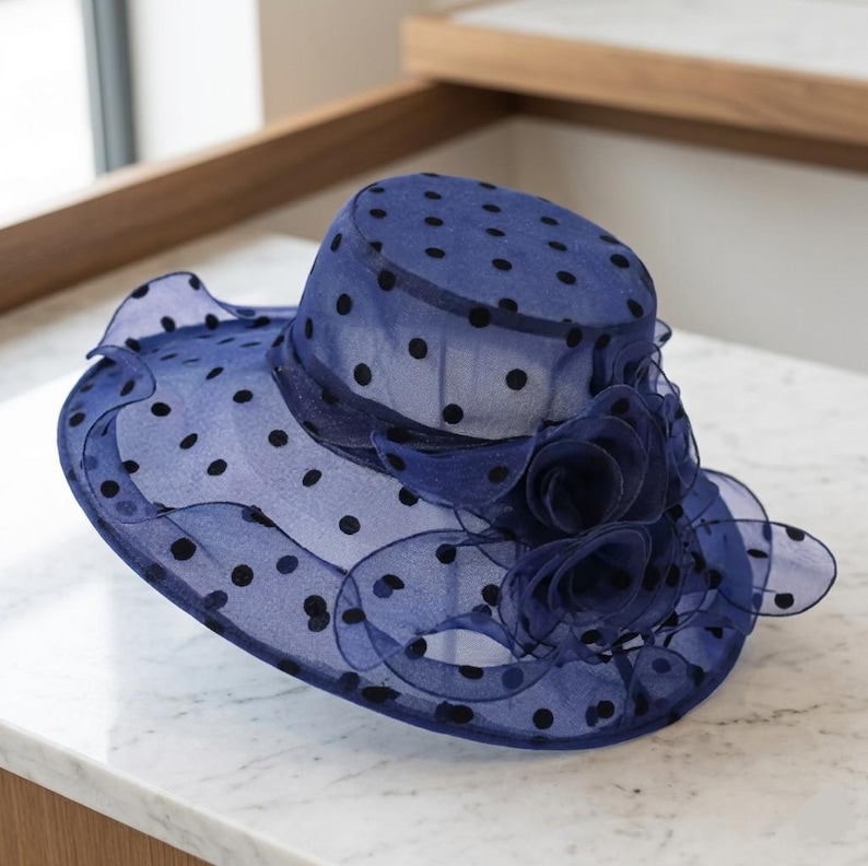 May include: A navy blue organza hat featuring a black polka dot pattern throughout the sheer fabric. The hat has a wide, wavy brim and is decorated with a cluster of layered, ruffled fabric flowers on the side of the crown and brim base.