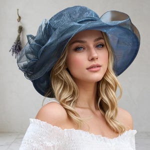 May include: A wide-brimmed hat in a deep slate blue hue, crafted from a shimmering, translucent organza-style fabric. The hat features a large, layered floral accent on the side with ruffled edges and a structured, slightly wavy brim design.