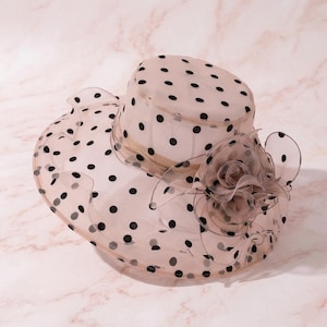 May include: A beige wide-brimmed hat made of sheer organza fabric featuring a black polka dot pattern throughout. The hat includes a structured crown and a large, layered organza flower embellishment on the side with ruffled edges.