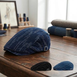 Könnte beinhalten: Eine dunkelblaue Jeans-Schiebermütze mit strukturiertem, diagonalem Streifenmuster und einer seitlichen Schnalle aus silberfarbenem Metall. Zwei kleinere Versionen der Mütze sind im Vordergrund zu sehen, eine in Schwarz und eine in hellblauem Jeansstoff.
