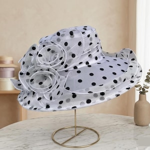 May include: A white wide-brimmed hat featuring a black polka dot pattern throughout the sheer fabric. The hat is adorned with two large, layered fabric rosettes on the side, creating a voluminous, ruffled appearance across the crown and brim.