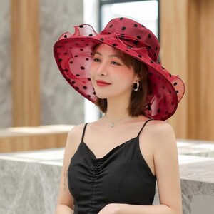 May include: A wide-brimmed hat in a deep red hue featuring a black polka dot pattern throughout the fabric. The hat is constructed from a sheer, layered material with a ruffled edge and a matching ribbon detail wrapped around the crown area.