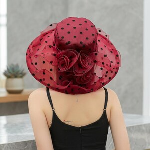 May include: A wide-brimmed hat in a deep burgundy red color featuring a sheer, translucent fabric with a black polka dot pattern. The hat is adorned with a cluster of layered fabric roses at the front and has a ruffled, wavy brim edge.