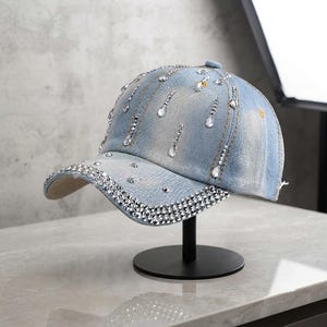 Puede incluir: Una gorra de béisbol de mezclilla azul claro con un acabado desgastado. La visera está decorada con filas de pequeños diamantes de imitación redondos y transparentes. La copa presenta cristales transparentes en forma de gota.