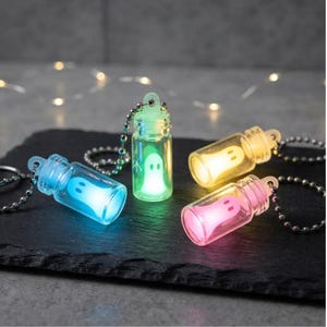 May include: Four small glass bottle keychains, each containing a glowing ghost figure. The ghosts emit blue, green, yellow, and pink light. Each clear glass vial is attached to a silver-toned metal ball chain, creating a set of illuminated charms.