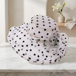 May include: A white wide-brimmed hat made of sheer, translucent fabric featuring a black polka dot pattern throughout. The hat includes a structured crown and a large, layered fabric flower accent on the side with ruffled, wavy edges.