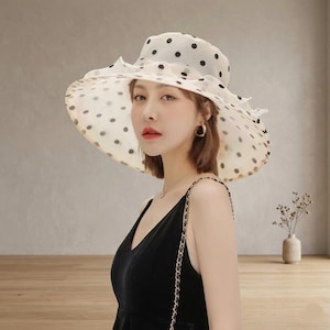 May include: A wide-brimmed hat made of sheer, off-white fabric featuring a black polka dot pattern. The hat has a structured crown and a ruffled, layered brim edge. The material appears lightweight and semi-transparent, creating a soft visual effect.