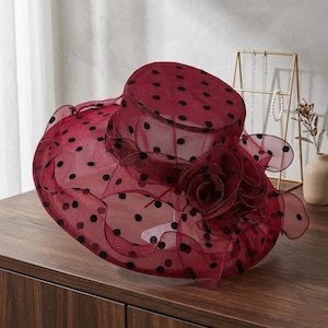 May include: A wide-brimmed hat made of sheer, dark red mesh fabric featuring a black polka dot pattern. The hat includes a structured crown and a ruffled brim, accented with a large, layered fabric flower detail on the side of the crown.
