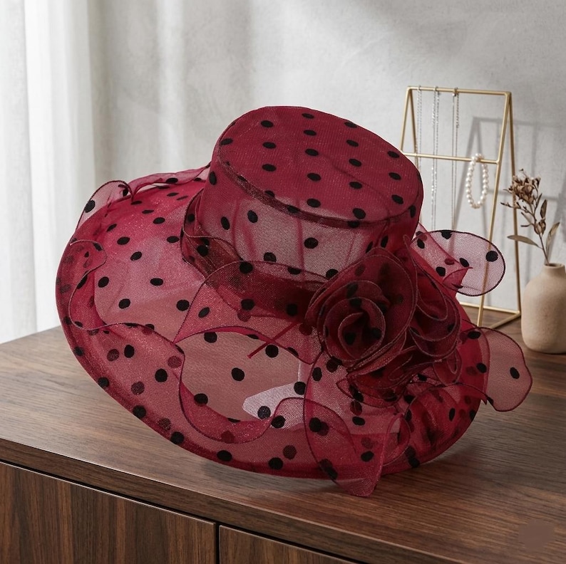 May include: A wide-brimmed hat made of sheer, dark red mesh fabric featuring a black polka dot pattern. The hat includes a structured crown and a ruffled brim, accented with a large, layered fabric flower detail on the side of the crown.