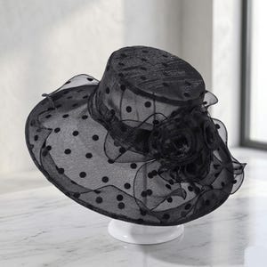 May include: A black sheer organza hat featuring a polka dot pattern throughout the crown and wide brim. The hat is adorned with a layered fabric flower detail on the side, constructed from matching black translucent material with ruffled edges.