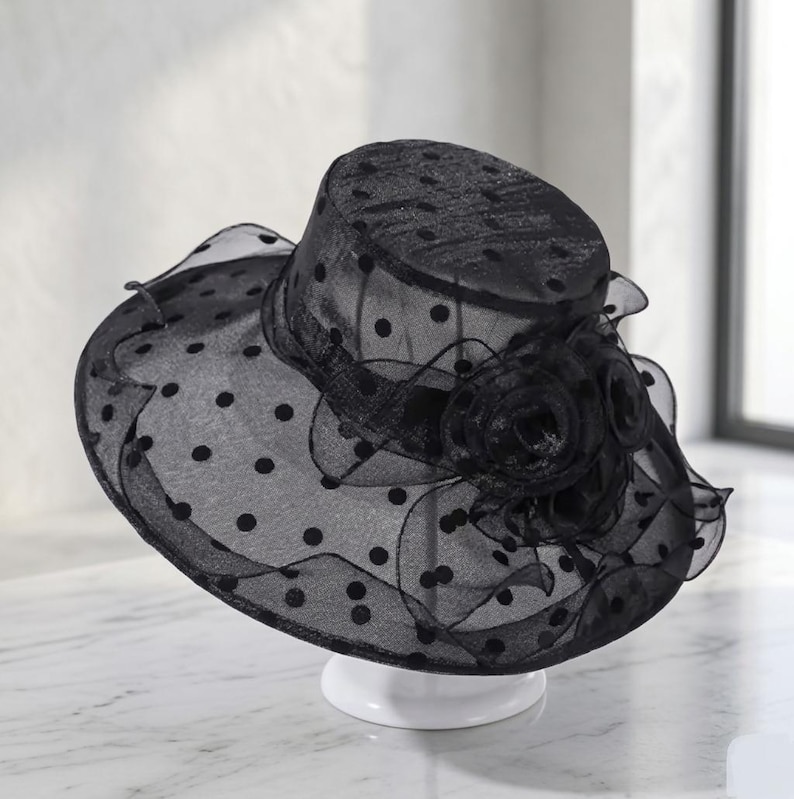 May include: A black sheer organza hat featuring a polka dot pattern throughout the crown and wide brim. The hat is adorned with a layered fabric flower detail on the side, constructed from matching black translucent material with ruffled edges.